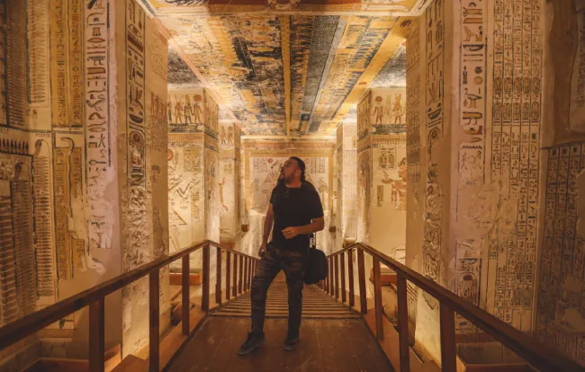 Interior view of the ancient Egyptian Tomb of Ramesses VI (KV9) in the Valley of the Kings, Luxor, featuring walls covered in detailed hieroglyphics and a highly decorated astronomical ceiling with a traveler standing on the wooden walkway looking up.